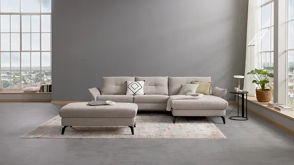 Modernes Wohnzimmer mit beigem Sofa, Hocker und buntem Teppich. Fenster bietet Blick auf Stadtlandschaft im Hintergrund.