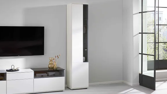 Nahaufnahme einer schmalen weißen Interliving Vitrine mit Glaselement neben einem TV-Lowboard.